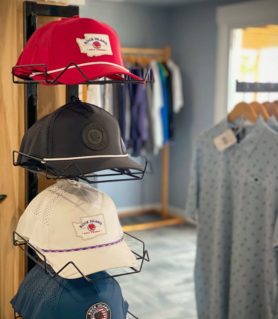 Caps on a wire rack: red, black, white, and blue, each with "Rock Island" logo. A light blue shirt with a small pattern hangs on a rack in the background.