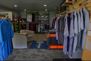 Golf shop interior with racks of colorful shirts on the right, a display of hats and pants in the back, two beige chairs on the left, and a branded mat on the floor.