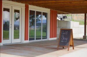 Outdoor view of a golf shop with a red and white exterior. A wooden sign with "Golf Shop" and an arrow points left.