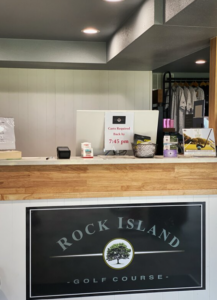 Reception desk at Rock Island Golf Course, featuring a wooden counter and a sign stating, "Carts Required Back by 7:45 pm." Shirts hang in the background.