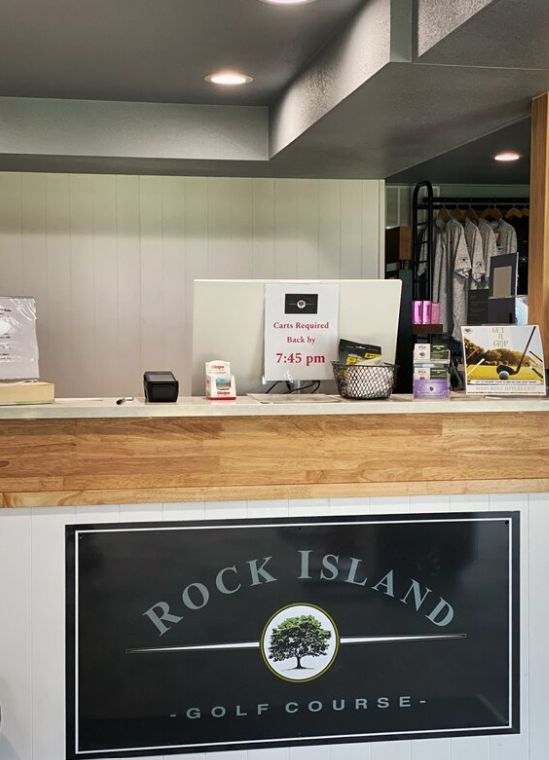 Reception desk at Rock Island Golf Course, featuring a wooden counter and a sign stating, "Carts Required Back by 7:45 pm." Shirts hang in the background.