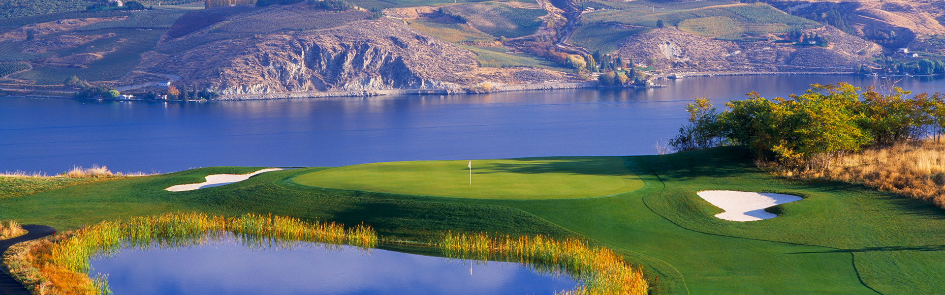 Scenic golf course beside a tranquil lake, with a vibrant green, sand bunkers, and autumn foliage. Rolling hills in the background under a clear sky.