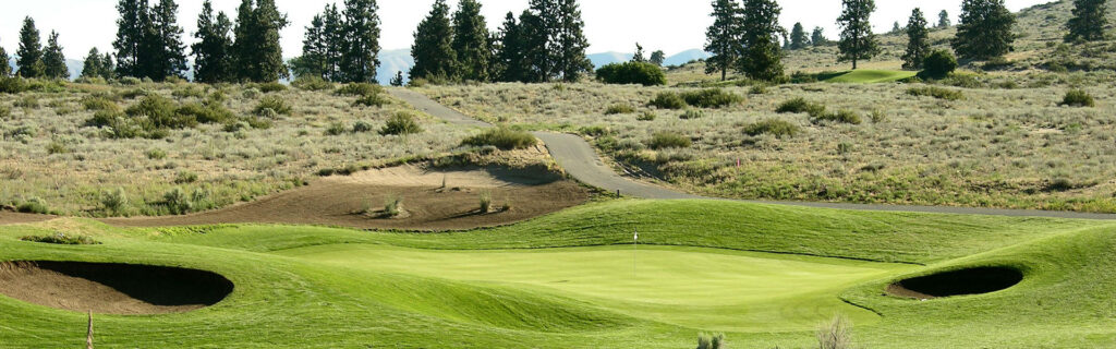 Golf course with a smooth, green fairway and two sand bunkers. A winding path leads through arid, hilly terrain dotted with shrubs and tall trees.