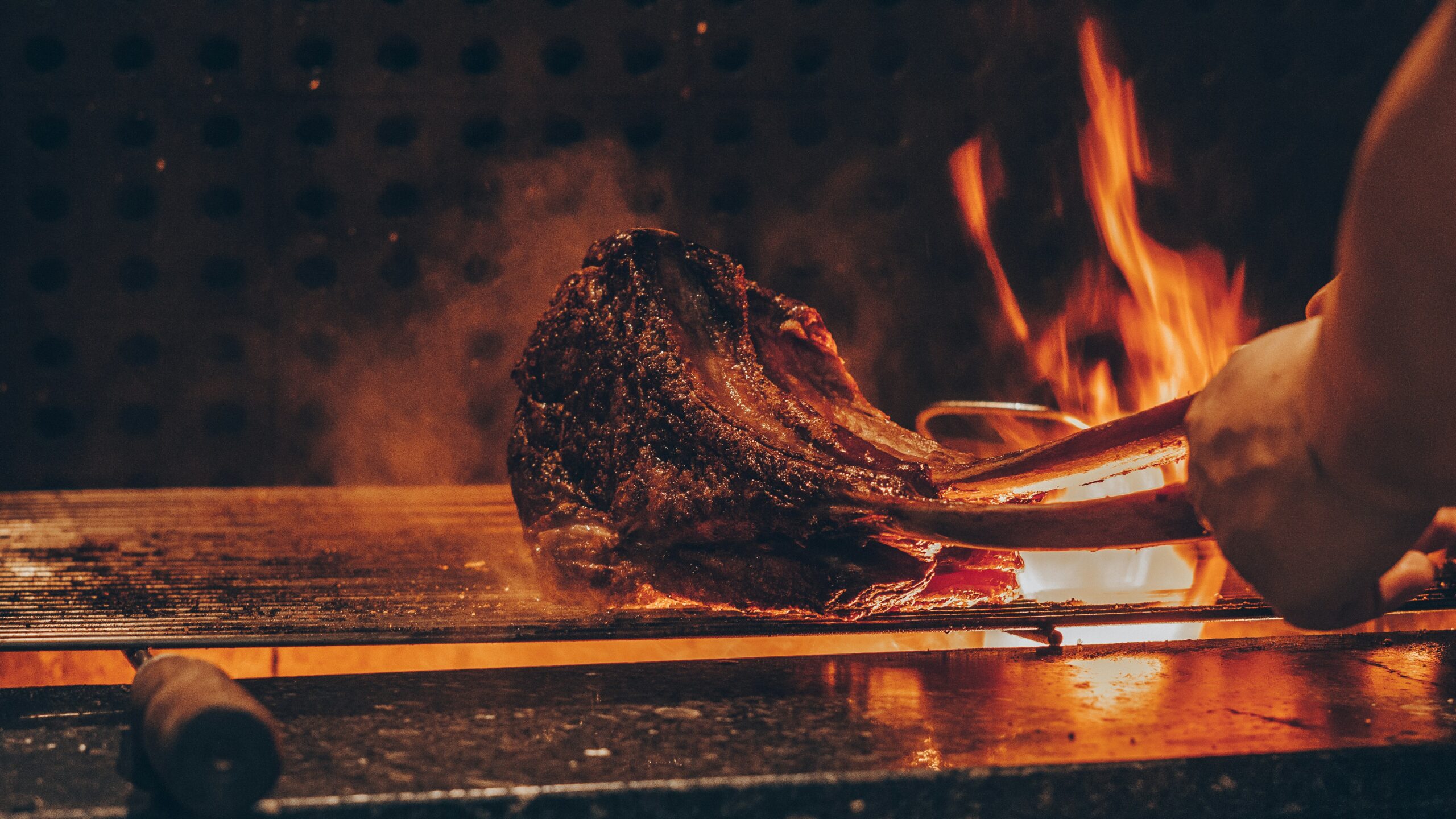 Steak is being grilled over an open flame. Tongs hold the meat as flames flicker in the background.