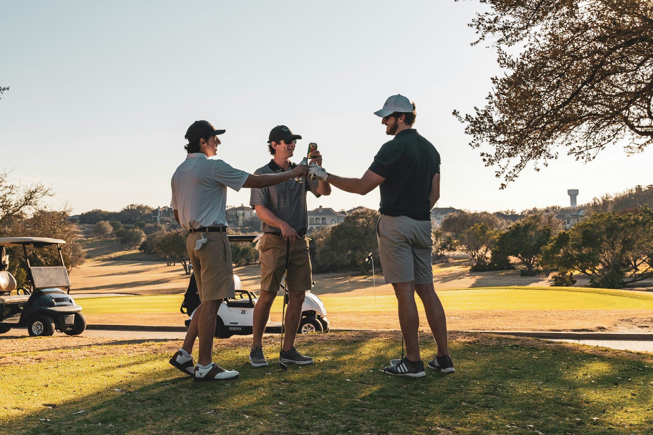 Three men in casual golf attire relax on a sunlit golf course, raising beer bottles in a cheerful toast, with a golf cart parked nearby.